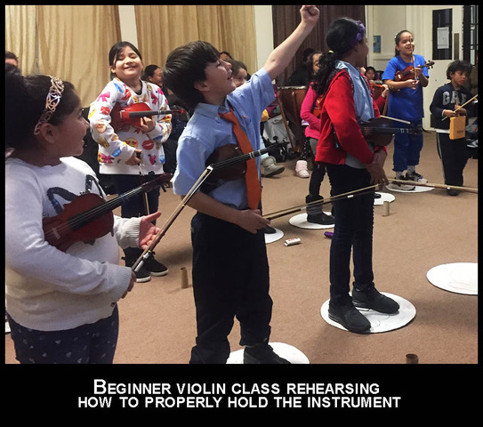 Image shows multiple young children with instruments in their hands at a beginner violin class rehearsing how to properly hold the instrument